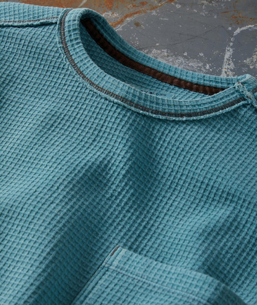 I Feel Better All Over, close-up of a blue textured long-sleeve shirt with a round neckline and chest pocket on a rustic wooden surface.