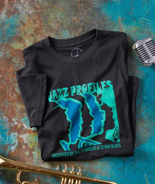 The Show Must Go On, black T-shirt with vibrant blue and turquoise jazz-themed graphic design and text, laid flat on a colorful textured surface, alongside a vintage microphone and a brass trumpet.