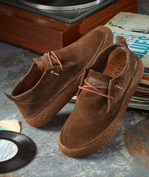 Kick Out, brown suede casual shoes with textured soles and laces, placed on a rustic surface next to vinyl records and magazines.