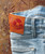 32 Bar Blues Take Me Back, close-up of light blue denim jeans with orange leather patch displaying the product name, vintage style background.