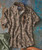 32 Bar Blues Funcalypso, a patterned short-sleeve button-up shirt with leaf designs, paired with sunglasses and green pants on a textured background.