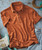Product 32 Bar Blues Who's Got Rhythm? , casual brown short-sleeve polo shirt with a collar, accompanied by a turquoise electric guitar, laying on a textured blue surface with guitar cables nearby.