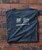 Product 32 Bar Blues Play It Forward, a folded dark gray T-shirt with white text and logos displayed against a brick wall background.