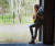 32 Bar Blues Jeff Fasano - Americana Portrait Sessions, a young woman playing an acoustic guitar outdoors beside a rustic building, with a background of green trees and bright natural light.
