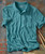 Upward Spiral, a turquoise textured polo shirt with a pocket and collar, laid flat on a rustic surface with drumsticks.