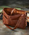 Somebody's Got To Go leather handbag, stylish brown leather tote bag with stitched detailing and adjustable strap, on rustic wooden surface with guitar in background.