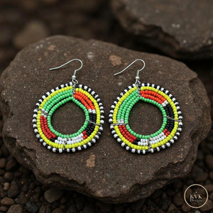 A pair of colorful beaded hoop earrings with green, yellow, red, white, and black beads arranged in concentric patterns, displayed on dark brown textured stones.