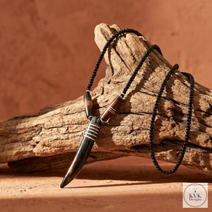 A beaded black necklace with a pendant shaped like a polished animal tooth, accented with silver bands, is displayed draped over a piece of weathered wood against a warm, earthy background.