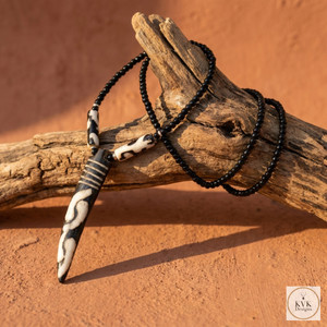 A black beaded necklace with a long, patterned pendant is draped over a piece of driftwood, set on a textured, sunlit surface. The image includes a small KVK Designs logo in the corner.