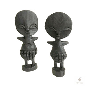 Two black fertility doll sculptures feature simplified human-like forms with large round heads and geometric bodies. The figures are standing on rectangular bases, both facing front.  The background is plain white.