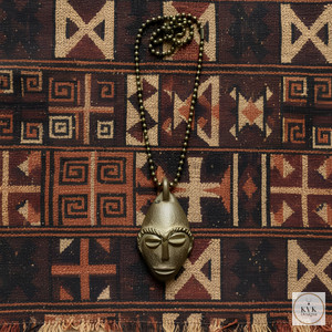 A beaded necklace with a metallic, stylized African mask pendant is laid on a woven fabric featuring geometric tribal patterns in earthy tones. The pendant is centered on the patterned textile background. A beaded necklace with a metallic, stylized African mask pendant is laid on a woven fabric featuring geometric tribal patterns in earthy tones. The pendant is centered on the patterned textile background.