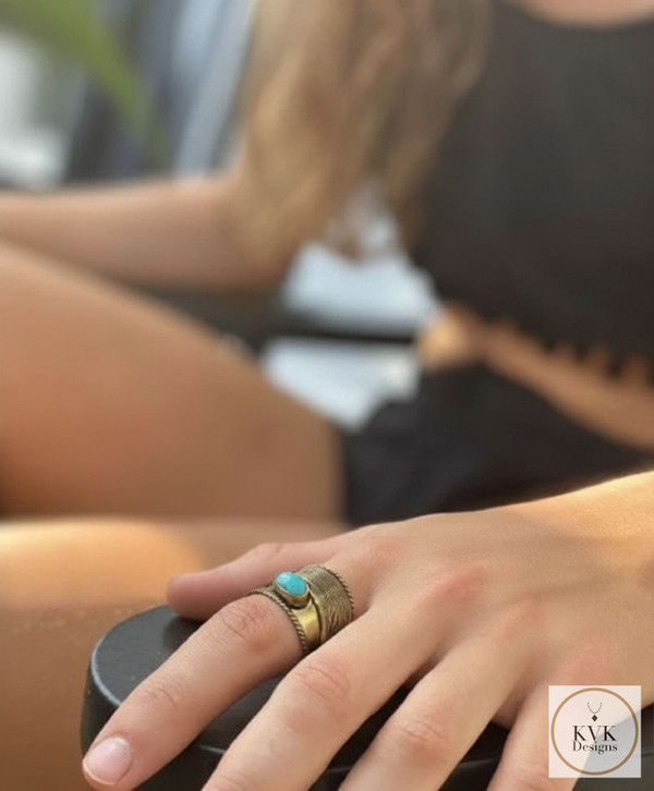 A woman's hand rests on a black surface, wearing a brass ring with a turquoise stone. She is dressed in a black outfit, and her long, wavy hair is partially visible. The KVK Designs logo appears in the lower right corner.