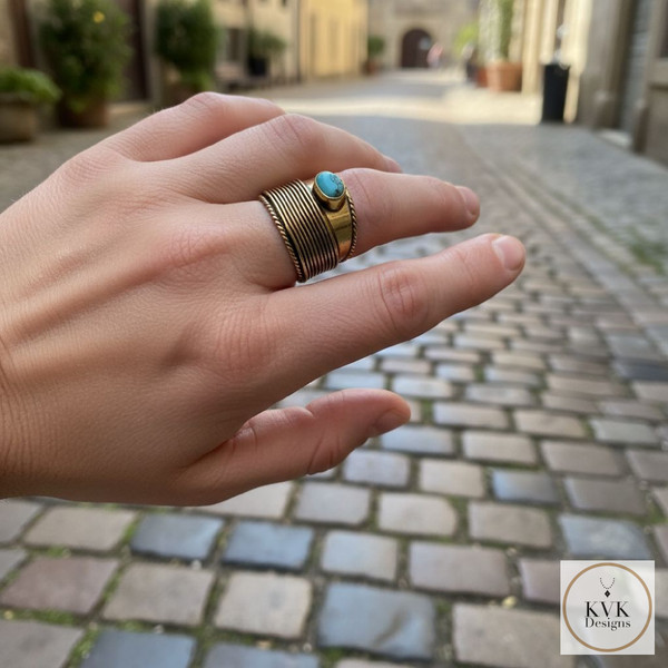 A hand wearing a brass ring with a turquoise stone is shown against a background of a cobblestone street in a European town. The logo "KVK Designs" appears in the bottom right corner.