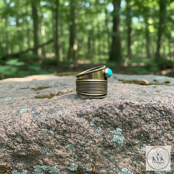 A bronze, ring with a turquoise stone sits on a large, mossy rock in a sunlit forest. The KVK Designs logo is visible in the bottom right corner.