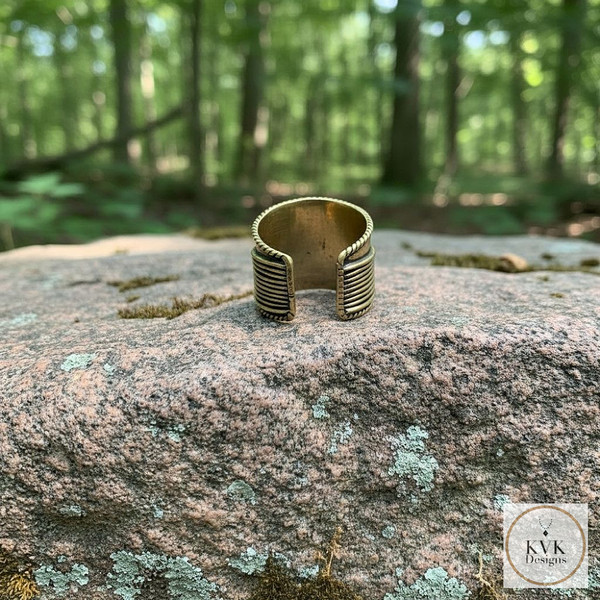 A brass adjustable ring with black striped detailing sits on a moss-covered rock in a sunlit forest. The KVK Designs logo appears in the bottom right corner.