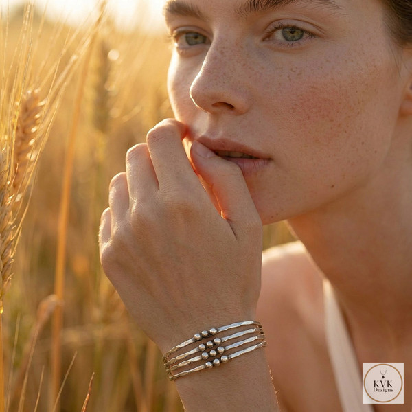 A woman with light skin and green eyes stands in a wheat field at sunset, resting her hand on her face and wearing a silver bracelet with multiple bands and bead accents. The KVK Designs logo is in the lower right corner.