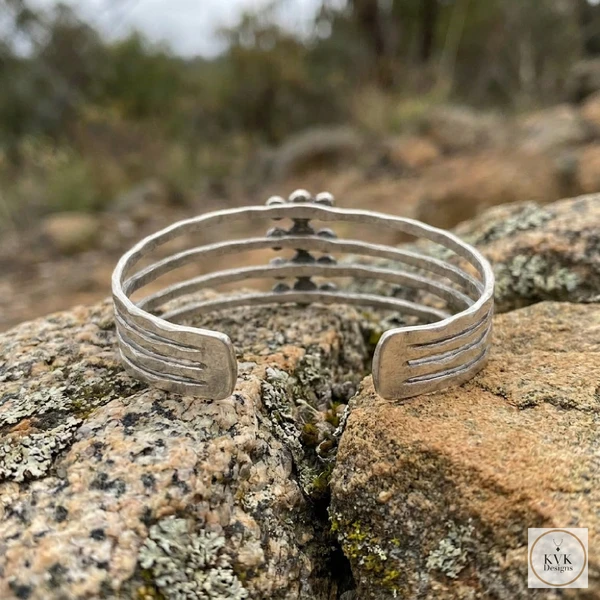 A handcrafted silver cuff bracelet with five parallel bands sits on a textured, lichen-covered rock outdoors, with a blurred natural background and a small KVK Designs logo in the corner.