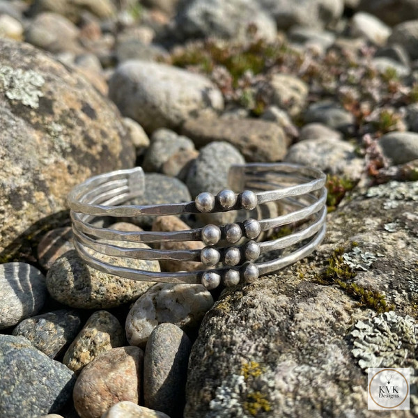 A silver cuff bracelet with five parallel bands and a cluster of metal beads in the center, displayed on a bed of natural rocks and pebbles.