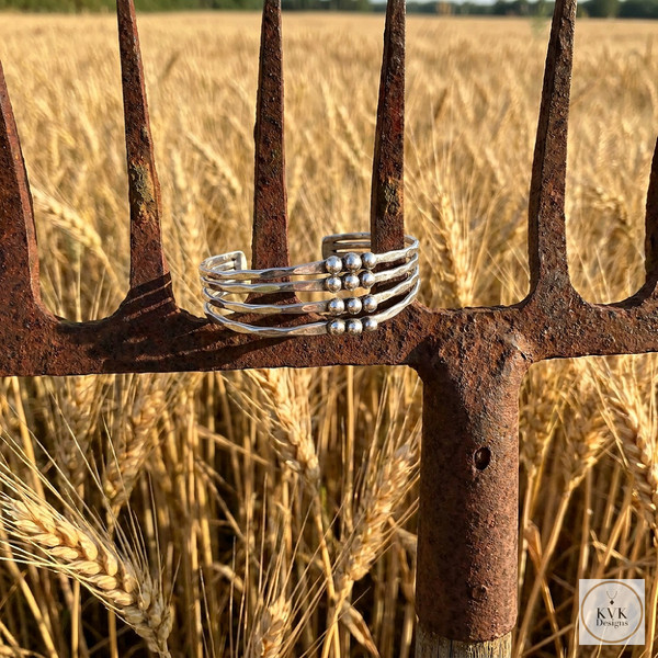A silver multi-band ring with small beads is displayed on the rusty prongs of a pitchfork, set against a background of a golden wheat field under a clear sky.