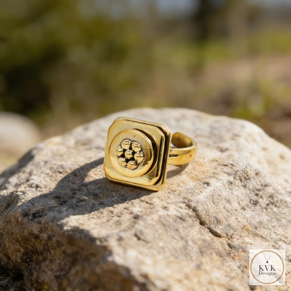 A gold ring with a raised square and floral design sits on a rough rock outdoors, with a blurred green background. The KVK Designs logo appears in the lower-right corner.