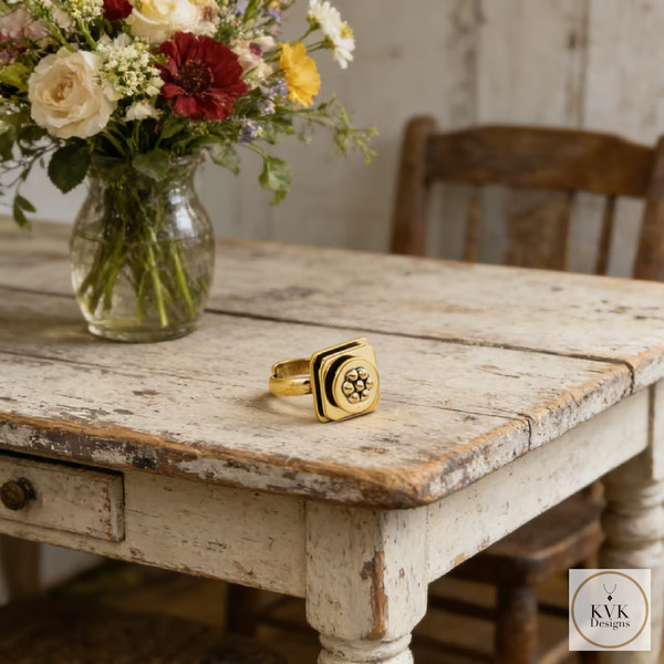 A gold ring with a flower design rests on a rustic, distressed wooden table next to a vase of colorful flowers; a wooden chair and a white wall are in the background. KVK Designs logo is in the bottom right corner.