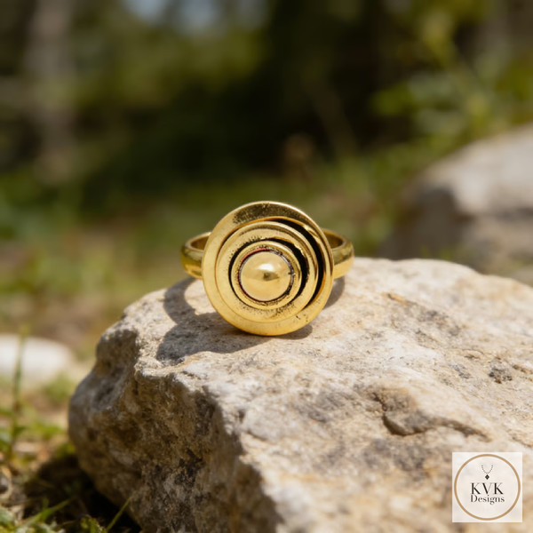 A gold ring with a circular, layered design sits on a large rock outdoors, with blurred greenery in the background. The KVK Designs logo is in the bottom right corner.