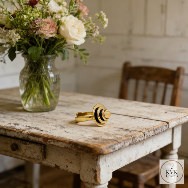 A gold ring with a modern design sits on a worn, rustic wooden table next to a glass vase filled with mixed flowers. An old wooden chair is in the background. The KVK Designs logo is in the bottom right corner.