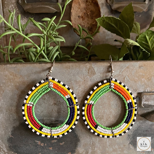 A pair of colorful, beaded hoop earrings featuring green, yellow, red, white, black, and blue beads, displayed hanging on a rustic metal container with green plants in the background.