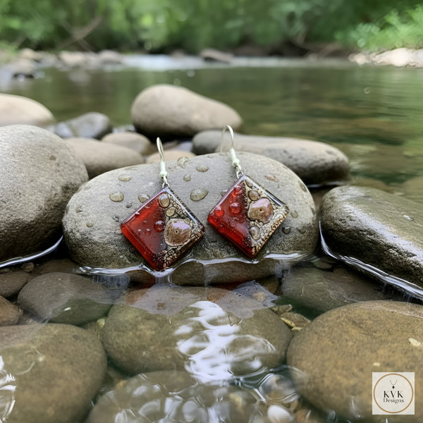 A pair of square, red and gold earrings rest on wet river stones in a shallow stream, with water and greenery in the background.