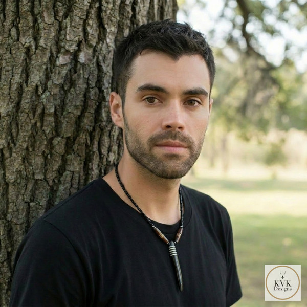 A man with short dark hair and a trimmed beard stands outdoors by a tree, wearing a black shirt and a necklace with beads and a tooth-shaped pendant. The background is blurred greenery. Logo for KVK Designs in the corner.