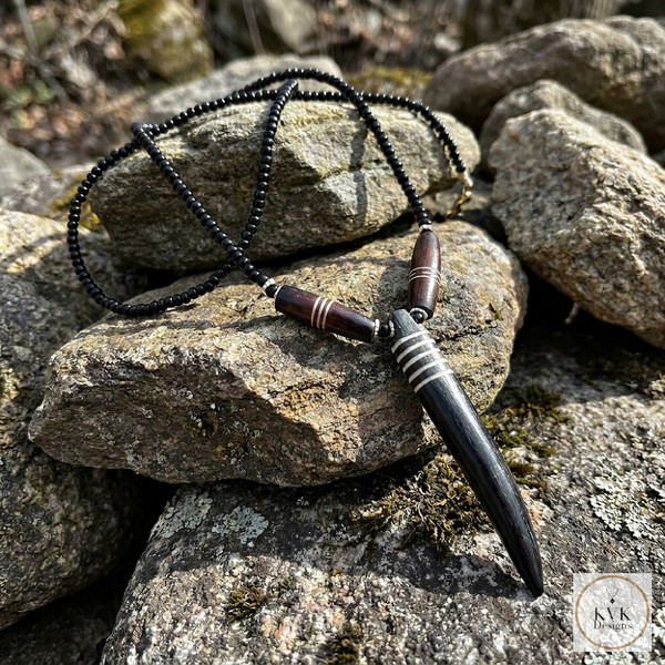 A black pendant necklace with silver accents and wooden beads is displayed on a pile of large, weathered rocks. Sunlight highlights the textures of both the stones and the jewelry. A logo reads "KVK Designs.
