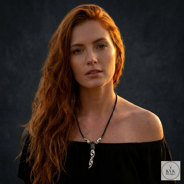 A woman with long red hair wears an off-shoulder black top and a black necklace with a white pendant, standing against a dark background. The logo "KVK Designs" is in the bottom right corner.