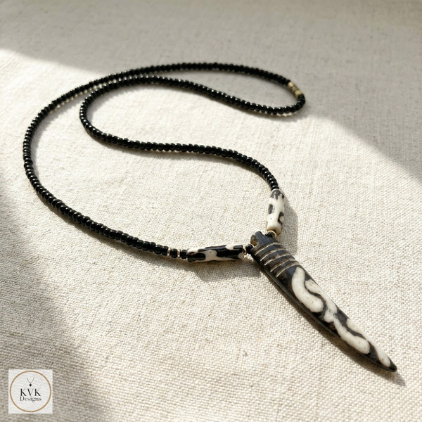 A black beaded necklace with a long, pointed, patterned pendant and matching bead detail is displayed on a light fabric surface. The KVK Designs logo is visible in the bottom left corner.
