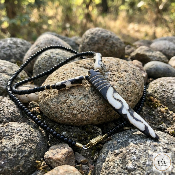 A black beaded necklace with a long, carved, black-and-white pendant rests on smooth rocks outdoors, surrounded by natural greenery and sunlight. A small logo in the corner reads "KVK Designs.