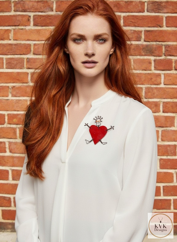 A woman with long red hair wears a white blouse with a large red heart brooch on the chest, standing in front of a brick wall. The brooch features a whimsical face and stick arms and legs. A KVK Designs logo is in the lower right corner.