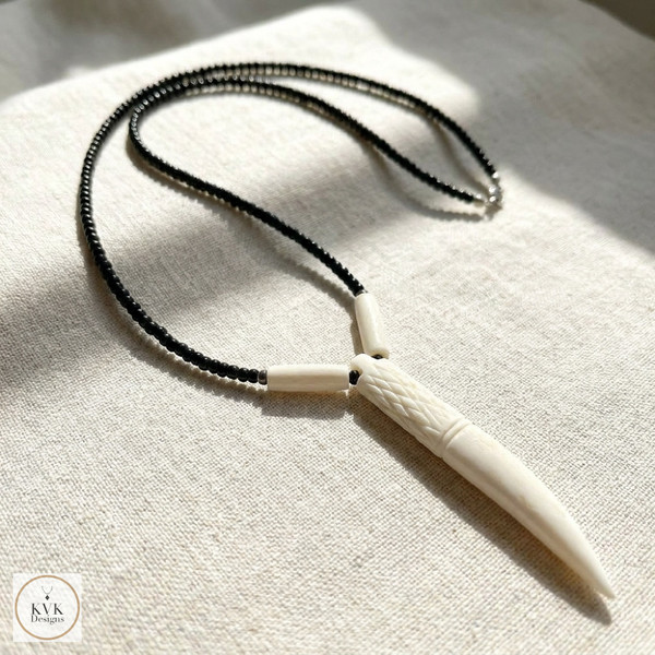 A beaded necklace with a carved ivory or bone pendant shaped like a fang or tusk, displayed on a light fabric surface with sunlight casting soft shadows. The necklace features black and white beads.