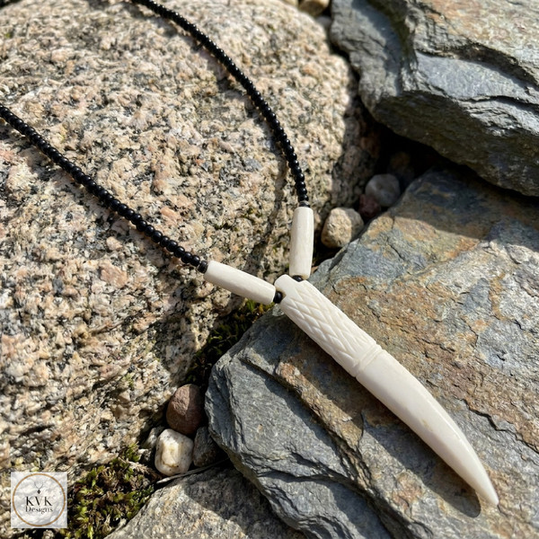 A beaded black necklace with a carved ivory or bone pendant lies on textured, layered rocks. The pendant has intricate details and is elongated, resembling a stylized fang or dagger. A small KVK Design logo is visible in the corner.