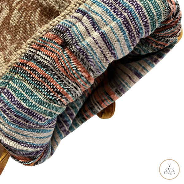Close-up of a colorful, striped woven fabric lining with shades of blue, orange, purple, and beige, folded at the edge. A tan strap and a small circular “KVK Designs” logo are visible in the corner.