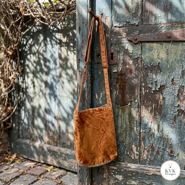 A small, yellow orange shoulder bag hangs on the handle of a weathered, blue-green wooden door with peeling paint. Sunlight casts shadows on the rustic scene. In the corner, a white circle shows the KVK Designs logo.