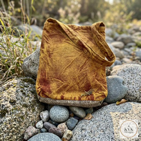 A yellow orange, tie-dye tote bag with a long strap and rests on smooth, gray river rocks outdoors. A circular “KvK Designs” logo appears in the bottom right corner.