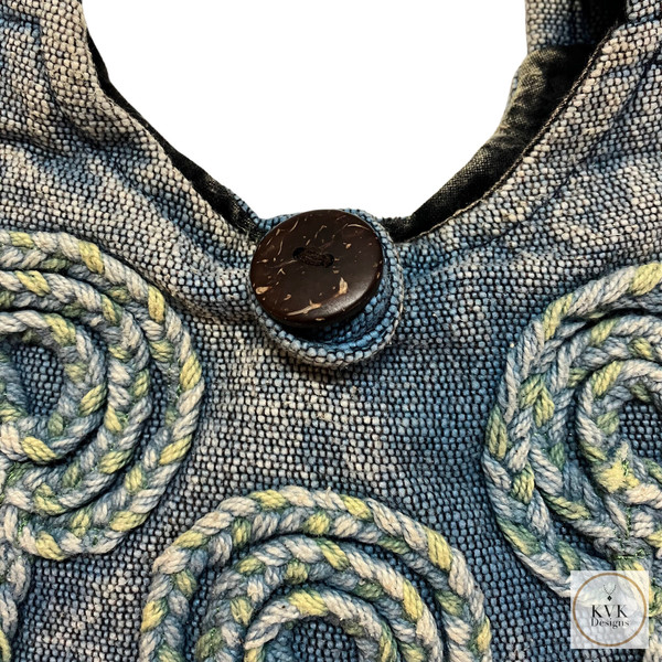 Close-up of a textured fabric bag with spiral embroidered designs in blue and green thread, featuring a round brown button closure at the top center. The KVK Designs logo is visible in the lower right corner.