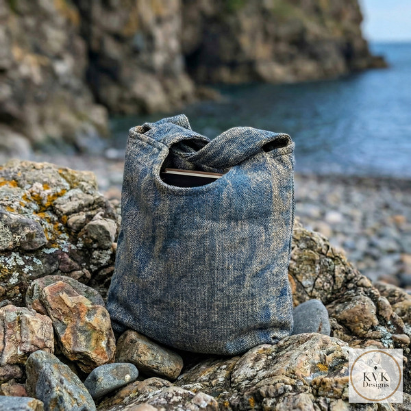A handmade blue fabric bag sits on rocky terrain by the seaside with cliffs and calm water in the background. The KVK Designs logo appears in the bottom right corner.
