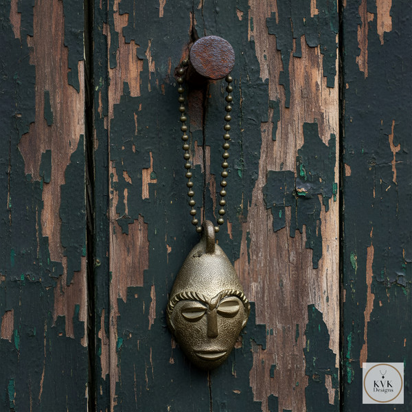 A bronze pendant shaped like a stylized human face hangs from a metal ball chain on a weathered, peeling wooden surface.