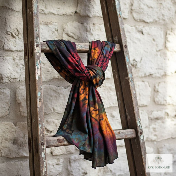 A colorful, tie-dyed scarf is loosely draped and knotted over the rung of a rustic wooden ladder, which leans against a white stone wall. The scene has a bohemian, artistic vibe.