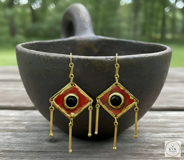 Pair of geometric gold and red earrings with dangling bars, hanging on the rim of a rustic black bowl, set on a wooden surface outdoors with greenery in the blurred background. Pair of geometric gold and red earrings with dangling bars, hanging on the rim of a rustic black bowl, set on a wooden surface outdoors with greenery in the blurred background.