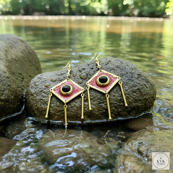 A pair of geometric gold earrings with black and pink details rest on a large rock in a shallow, flowing river with green trees blurred in the background. A pair of geometric gold earrings with black and pink details rest on a large rock in a shallow, flowing river with green trees blurred in the background.