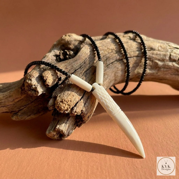 A carved ivory-colored pendant resembling a tooth or fang hangs from a black beaded necklace, draped over a weathered piece of driftwood on a peach-colored surface.