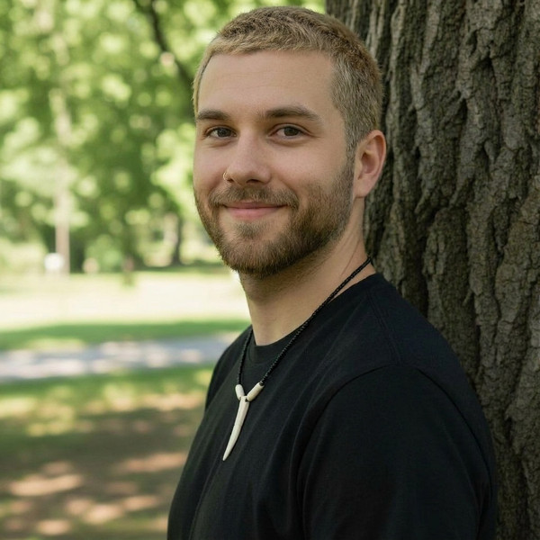 A young man with short hair and a beard, wearing a black shirt and a necklace with a white pendant, stands outdoors leaning against a tree, smiling, with a blurred green park background.