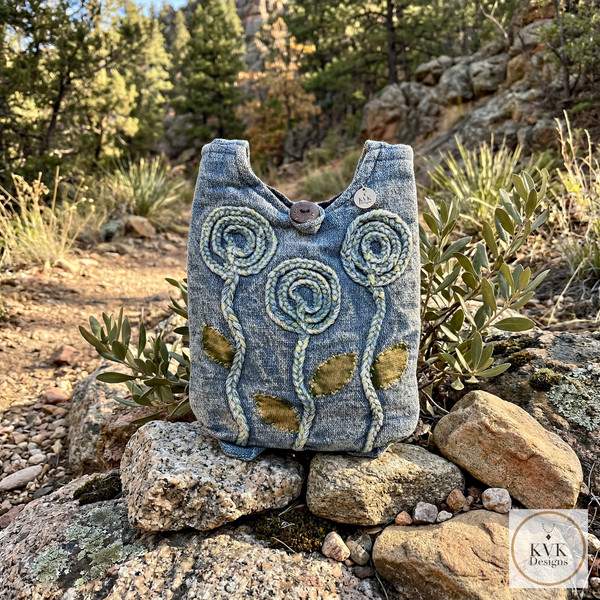 A handmade blue fabric bag with three embroidered flowers and green leaves sits on rocks in an outdoor, mountainous area with trees and brush. A circular "KvK Designs" logo is in the bottom right corner.