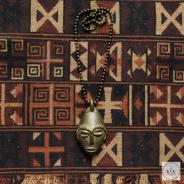 A beaded necklace with a metallic, stylized African mask pendant is laid on a woven fabric featuring geometric tribal patterns in earthy tones. The pendant is centered on the patterned textile background.